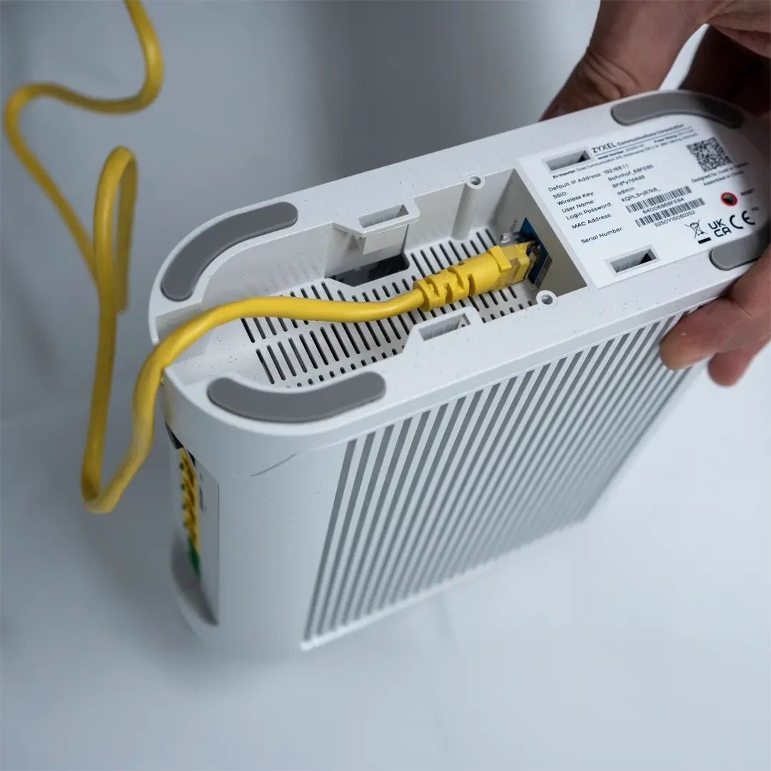 Henkilö pitää kädessään Zyxel-reititintä, johon on liitetty keltainen verkkokaapeli laitteen pohjassa olevaan porttiin.