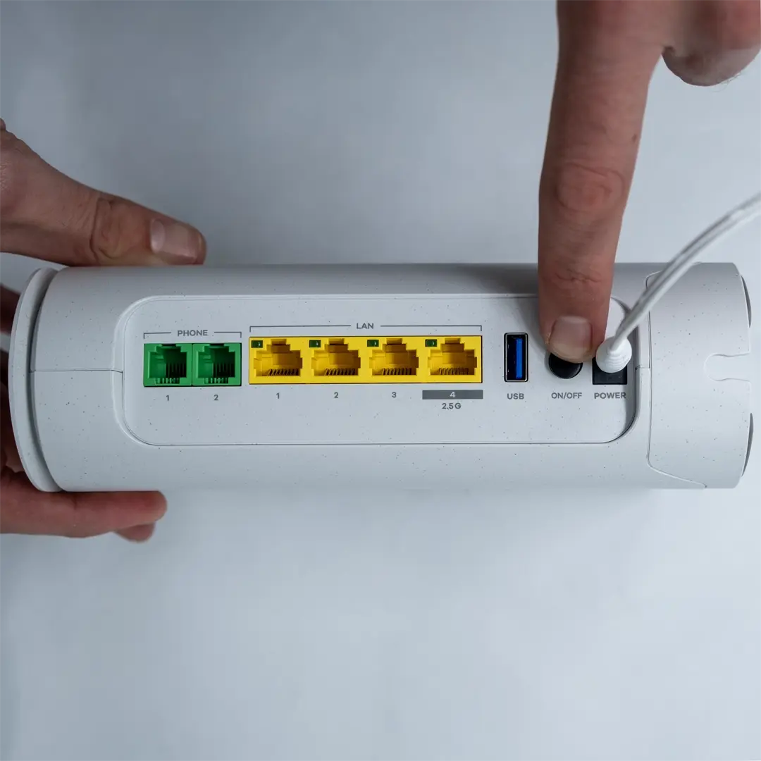 Henkilö painaa virtapainiketta Zyxelin reitittimen takana käynnistääkseen sen. Virtajohto on liitetty, ja keltaiset LAN-portit sekä USB-portti näkyvät selvästi.